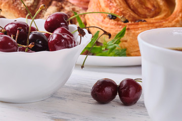 puffs with cherries on a white plate with a bowl of fresh cherries and a cup of tea on a white wooden background. Close-up. Selective focus