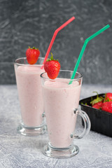 Two glasses with strawberry milkshake with pieces of berries on the edge of the glass on gray background.