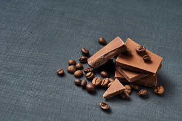 Heap of broken pieces of bitter or milk chocolate with fried coffee beans scattered on dark scratched concrete table. Space for text