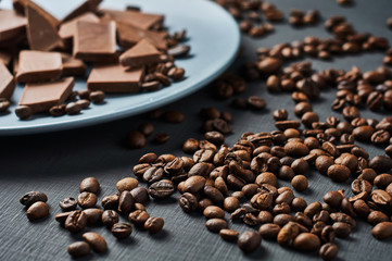 Heap of many broken pieces of bitter or milk chocolate on blue circle ceramic plate and scattered fried coffee beans lies on dark scratched concrete table. Close-up