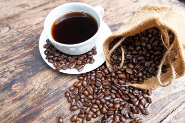 full of coffee beans spilling out bag on brown wooden background with a cup of black coffee