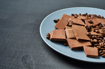 Heap of many broken pieces of bitter or milk chocolate and fried coffee beans on blue circle ceramic plate lies on dark scratched concrete table. Space for text