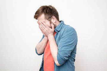 Man in a red t-shirt and blue shirt covers his face with his hands, on a white background.