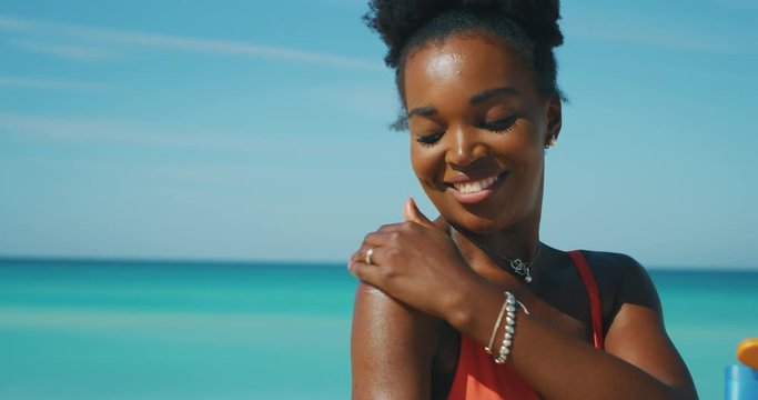 Slow motion of happy young african woman is applying a sunscreen or sun tanning lotion to take care of her skin during a vacation on a beach and smiling in camera.