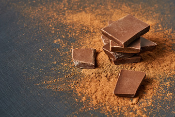 Heap of broken pieces of bitter or milk chocolate powdered of cocoa scattered on dark scratched concrete table. Space for text