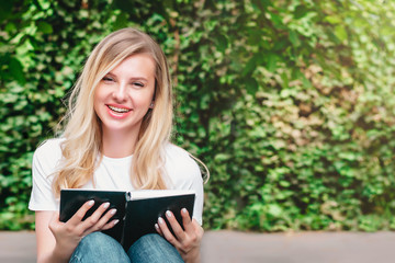 Fototapeta premium Young blonde girl sits on a bench, reads a book and smiles in a park on a background of trees and bushes