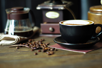 Closeup hot cappuccino cup with selective focus on coffee beans on wooden table with blurred grinder background