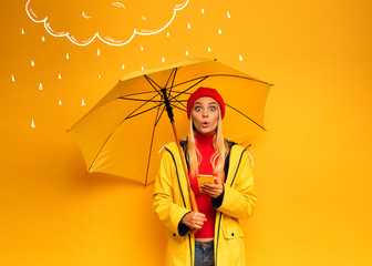 Girl with smartphone and umbrella on yellow background surprised for the weather - obrazy, fototapety, plakaty
