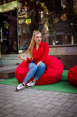 A beautiful girl of European race with very long and blond hair in a red shirt and jeans, sits on a red modern armchair in the city center. Model with professional makeup. The girl has beautiful lips 