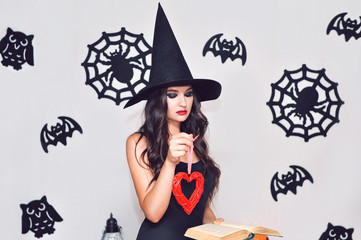 A young woman in a black witch hat does a voodoo ceremony with a red wicker heart and a needle.