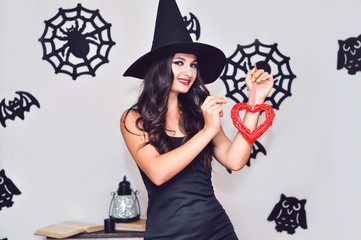 A young woman in a black witch hat does a voodoo ceremony with a red wicker heart and a needle.