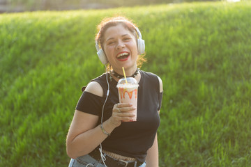 Beautiful sensual young woman listens to music with headphones and holds a milkshake in her hands while walking through the park on a sunny evening. Weekend getaway concept.