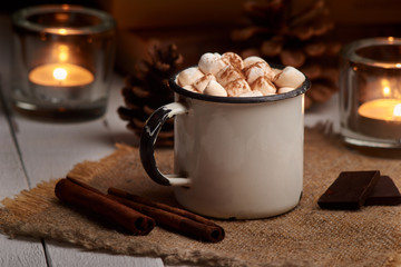 Cup of hot cocoa or hot chocolate with marshmallows and cinnamon sticks on wooden background with burning candles. Rustic. Winter mood.
