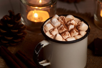 Cup of hot cocoa or hot chocolate with marshmallows and cinnamon sticks on wooden background with burning candles. Rustic. Winter mood.