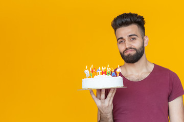 Positive funny young guy with a cap and a burning candle and a homemade cake in his hands posing on a yellow background. Anniversary and birthday concept.