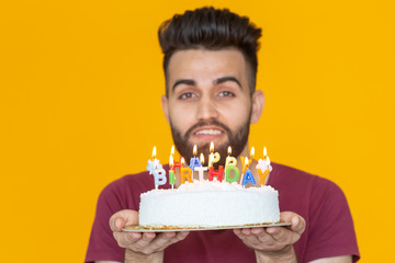 Close-up young handsome man blows off a candle from a burning cake posing for a yellow background. Holiday concept.