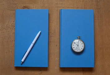 Two blue books, white pen and vintage pocket watch on wooden table background; top view, still life.