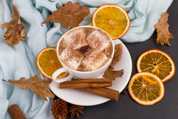 Cup coffee with marshmallows, cocoa, scarf, leaves, dried oranges, spices, on gray background. Delicious hot autumn drink, morning mood. Copy space.