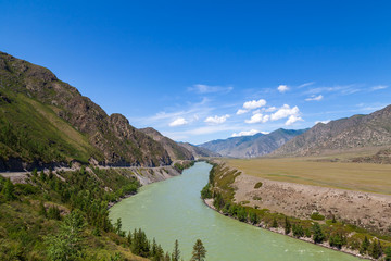 Fototapeta na wymiar Landscape of the mountain chain of the Altai covered with green trees and rocks, with the turquoise Katun River and its rapids on a sunny summer day and a blue sky with white clouds. Tourist route.