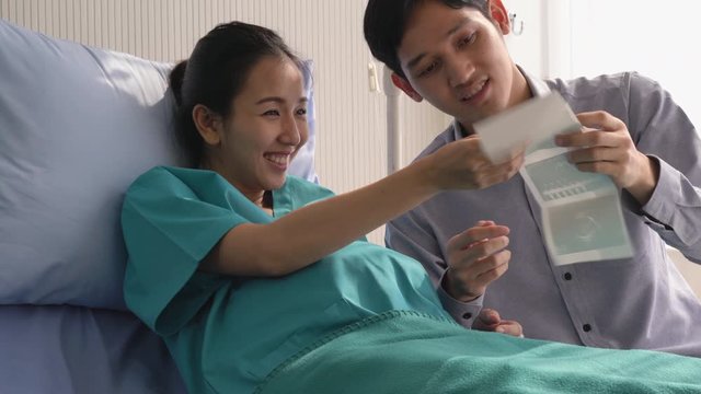 Asian family husband looking picture ultrasound scanning with wife happy emotion. Pregnant woman belly at hospital on the bed. Concept of family, hope and new born.