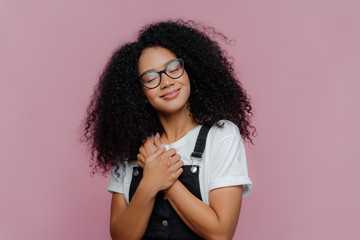 Portrait of satisfied woman with Afro hairstyle, keeps hands pressed on chest, tilts head, wears...