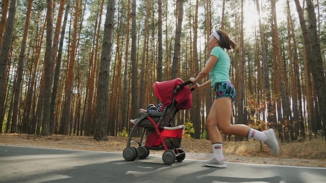 Running American girl with baby stroller enjoying a sunny summer day in a pine forest. Jogging or power walking, active family with baby jogger. Slow-motion 4k video