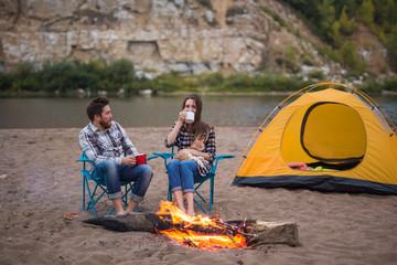 people, summer tourism and nature concept - couple near camp fire warming up seen from the tent