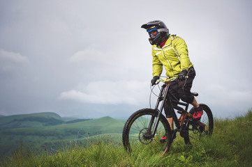 Fototapeta premium Front view of a man on a mountain bike standing on a rocky terrain and looking down against a gray sky. The concept of a mountain bike and mtb downhill