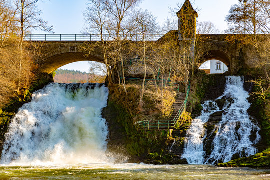 Rural stone vehicular bridge surrounded by leafless trees with a tower and two small waterfalls with abundant water flow on the Ambleve river, sunny day in the small town of Coo, Belgium
