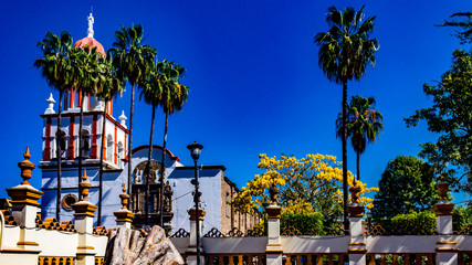 Exterior facade of the church of San Pedro Apostol surrounded by a white wall with yellow details and palm trees, wonderful sunny day in Tlaquepaque Jalisco Mexico