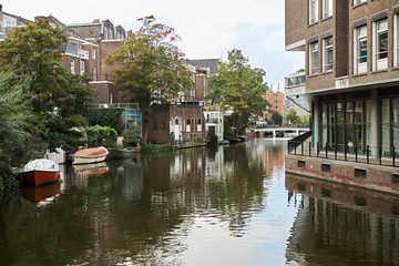 Fototapeta premium Artificial waterway, transportation, navigation, city landscape and tourism concept. Picture of channel running along urban buildings through European town. Boats on canal in cityscape Amsterdam