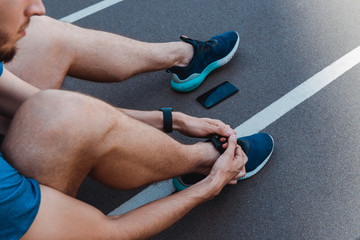 cropped view of young sportsman tying shoelaces near smartphone with blank screen on running track