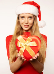 Guess what’s in the box! Cheerful young woman in a red dress and Santa hat with long blonde hair smiles and poses with a red heart-shaped box with a golden ribbon in her hands.