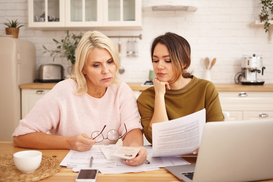 Picture of friendly caring young dark haired woman helping mother to fill in financial papers at home. Attractive blonde middle aged female is taught to pay utility bills online using laptop