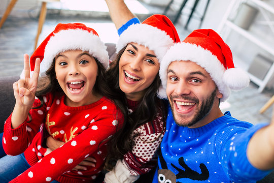 Eternal love. Close-up photo of young mom and dad hugging their little brunette daughter and laughing while taking a selfie in Santa caps at home on a Christmas day