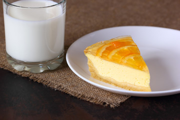 Cheesecake with oranges on a white plate, glass with milk on burlap tablecloth.