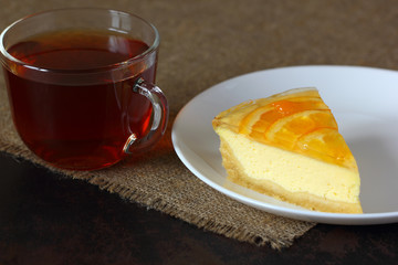 Cheesecake with oranges on a white plate, glass mug with tea on burlap tablecloth.