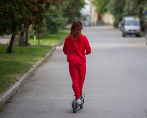 Red-haired girl in a red tracksuit drives an electric scooter. A young woman in oversized clothes rides around the city and listens to music using wireless headphones. View from the back.