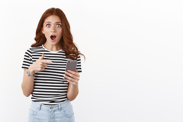 Shocked and amazed redhead girl in striped t-shirt, gasping with awe and amazement, pointing smartphone display, drop jaw stare camera astonished, gaze with awe and shock, stand white background