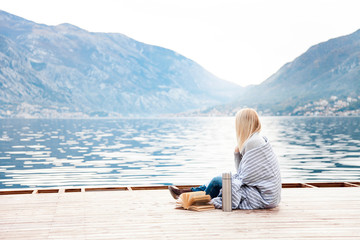 Woman on wooden pier by winter sea, mountains. Cozy picnic with coffee, hot beverages, tea in thermos and mug, warm plaid, opened book. Girl is enjoying nature, relaxation, reading on beach