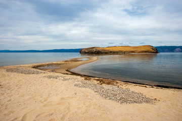 The sandy shore of Lake Baikal overlooking the sand spit and the island. High mountains are visible on the horizon. The sky with clouds. The weather is calm.
