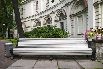 White wooden bench in an old city park. Nearby are green bushes and beautiful flowers.