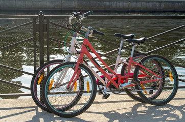 Fototapeta na wymiar In the midday two bikes white and red of travelling together persons parked and locked with anti-theft cables to the fence street on the background of azure water in the river