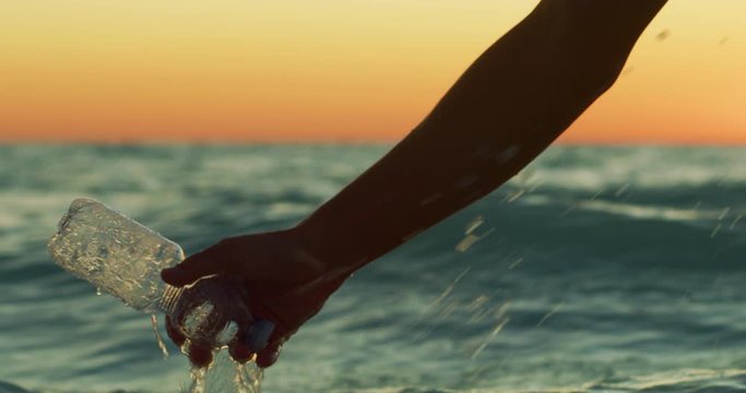 Slow motion close up of a hand of young volunteer is picking up a plastic bottle from a sea to protect an environment on a sunset.