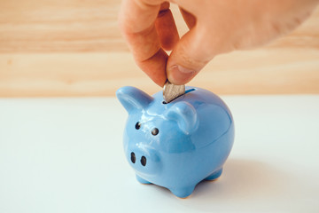Hand is putting coin in blue piggy bank on wooden and white background. © gesrey
