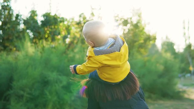 Mom plays with her child in the park at sunset. The boy sits on his shoulders in a yellow jacket and smiles happily. Spin together.