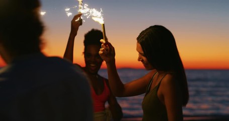 Slow motion close up of young multi-ethnic carefree friends are having fun to party with sparklers on a beach with a sea in twilight sunset.