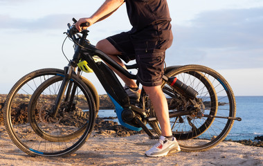 Fototapeta premium Senior man riding his bici on the cliffs. Horizon over sea at sunset light. Two electric bicycles with battery and engine. One people in activity