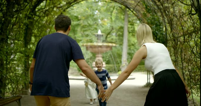 The family walks under the plant arch in the Park: a blonde mother catches her daughter with open arms, and a dark-haired father catches his son with his arms. They laugh.