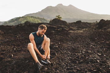 Young man sitting and tying running shoes before trail running outdoors. Volcanic landscape and...
